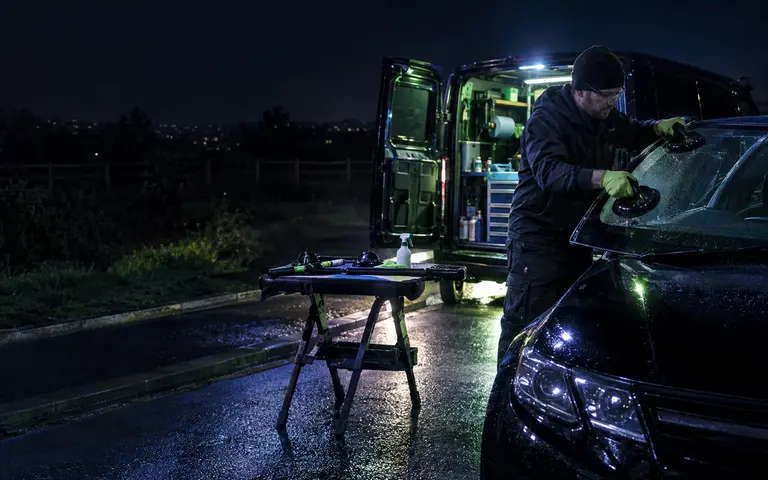 Technician working on a car windscreen beside a lit mobile service van at night